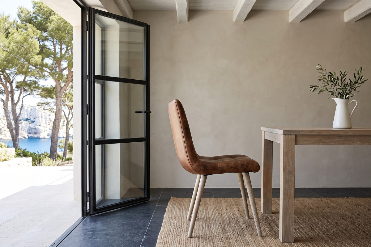 A Compiano dining chair in distressed brown leather in side profile in a Cassis villa, beside a bleached-elm table