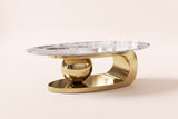 The Cosima coffee table, front three-quarter view: an oval sintered stone top with bold brown and charcoal veining on a polished gold base of a plinth, a large sphere and an open loop, on an off-white studio background