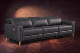 The Elodie sofa on a dark walnut herringbone floor against a plaster wall, three-quarter view, headrests down and footrests closed. Shown in Aged Slate, 3 seat.