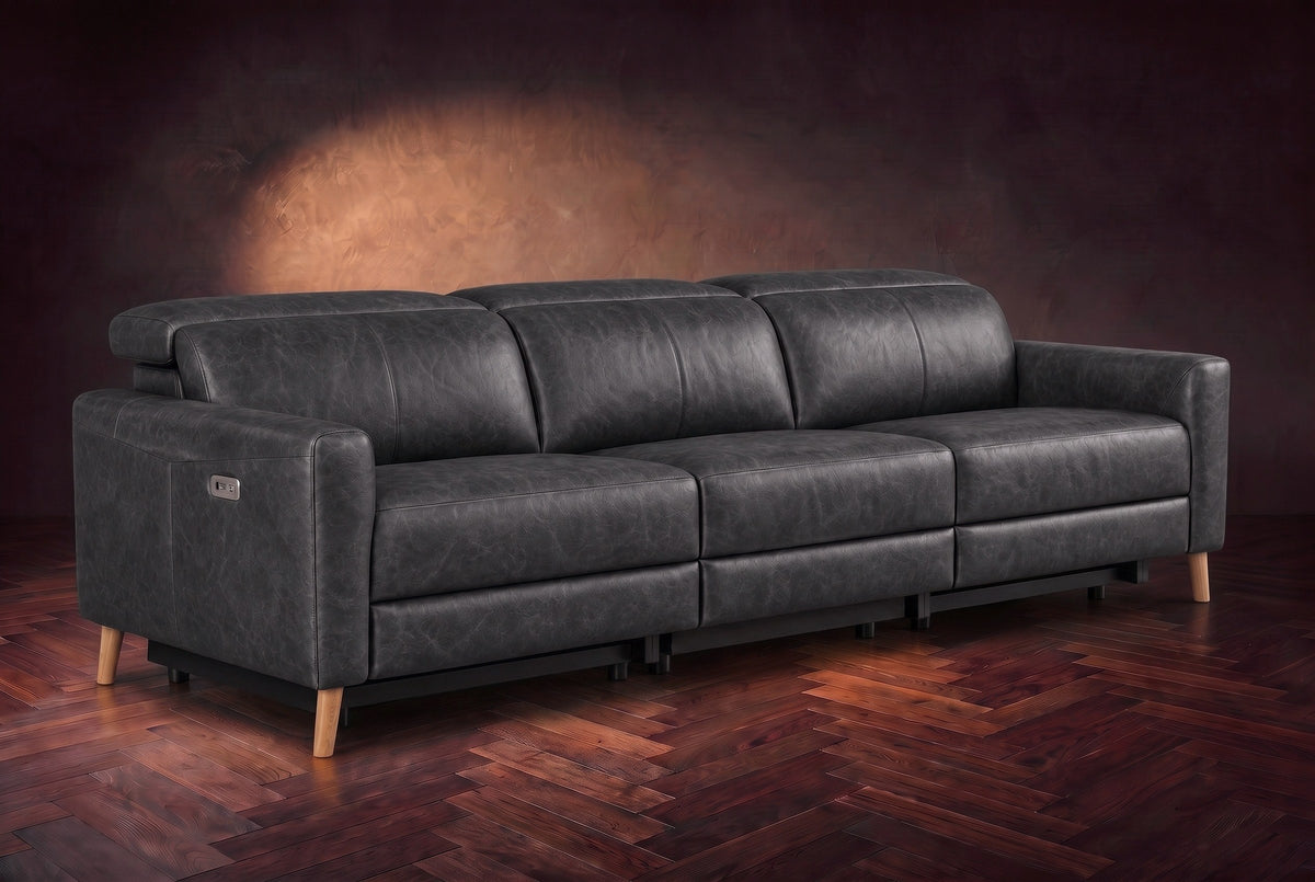 The Elodie sofa on a dark walnut herringbone floor against a plaster wall, three-quarter view, headrests down and footrests closed. Shown in Aged Slate, 3 seat.