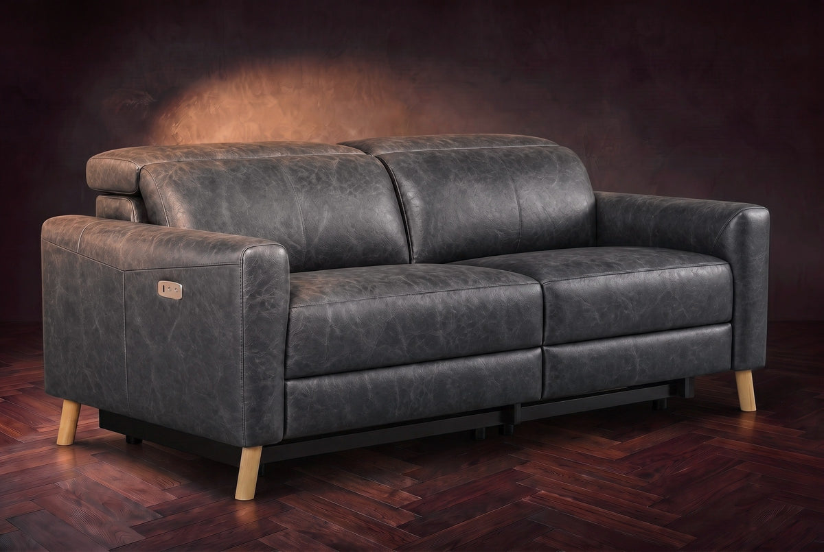The Elodie loveseat on a dark walnut herringbone floor against a plaster wall, three-quarter view, headrests down and footrests closed. Shown in Aged Slate, Loveseat.