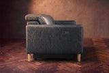 The left end of the Elodie sofa seen from the side on a dark walnut herringbone floor, the control panel on the arm and the headrest raised. Shown in Aged Slate, 3 seat.
