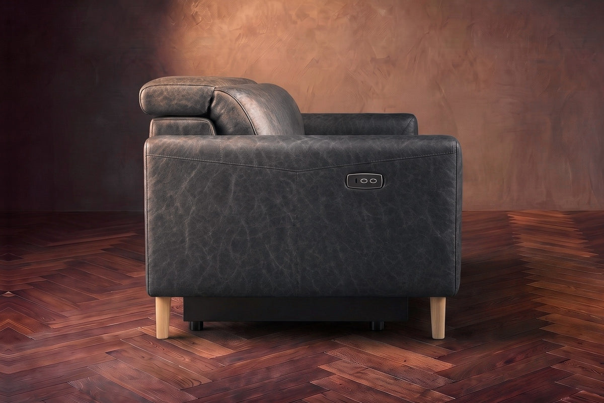 The left end of the Elodie sofa seen from the side on a dark walnut herringbone floor, the control panel on the arm and the headrest raised. Shown in Aged Slate, 3 seat.
