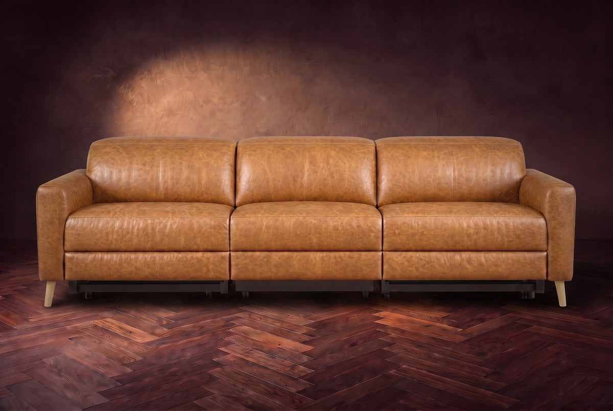 The Elodie sofa seen from the front on a dark walnut herringbone floor against a plaster wall, headrests down and footrests closed. Shown in Antiqued Caramel, 3 seat.