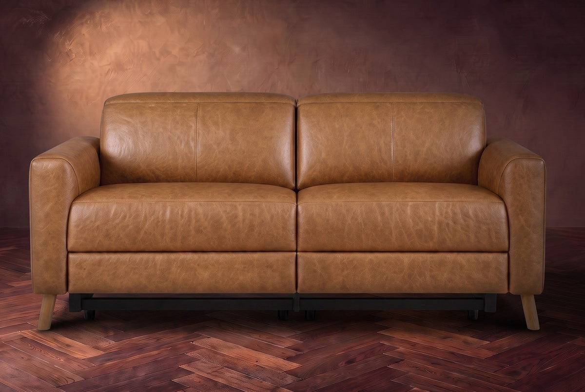 The Elodie loveseat seen from the front on a dark walnut herringbone floor against a plaster wall, headrests down and footrests closed. Shown in Antiqued Caramel, Loveseat.