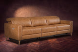 The Elodie sofa on a dark walnut herringbone floor against a plaster wall, three-quarter view, headrests down and footrests closed. Shown in Antiqued Caramel, 3 seat.