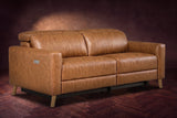 The Elodie loveseat on a dark walnut herringbone floor against a plaster wall, three-quarter view, headrests down and footrests closed. Shown in Antiqued Caramel, Loveseat.