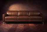 The Elodie sofa seen from the front on a dark walnut herringbone floor against a plaster wall, headrests down and footrests closed. Shown in Burnished Chestnut, 3 seat.