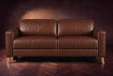 The Elodie loveseat seen from the front on a dark walnut herringbone floor against a plaster wall, headrests down and footrests closed. Shown in Burnished Chestnut, Loveseat.