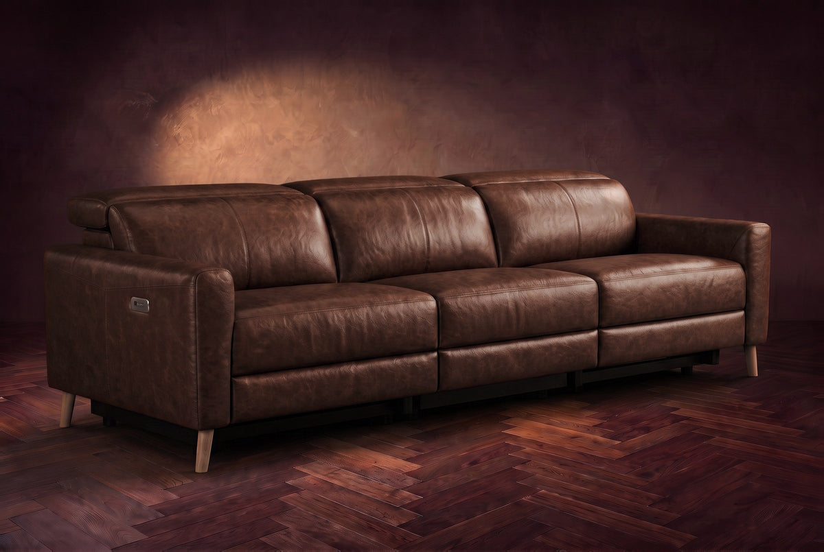 The Elodie sofa on a dark walnut herringbone floor against a plaster wall, three-quarter view, headrests down and footrests closed. Shown in Burnished Chestnut, 3 seat.