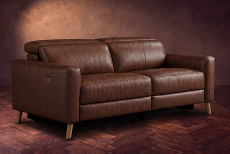 The Elodie loveseat on a dark walnut herringbone floor against a plaster wall, three-quarter view, headrests down and footrests closed. Shown in Burnished Chestnut, Loveseat.