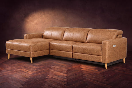 The Elodie sectional on a dark walnut herringbone floor in front of a warm plaster wall, three-quarter view, headrests down and the footrest closed. Shown in Antiqued Caramel, Left chaise.