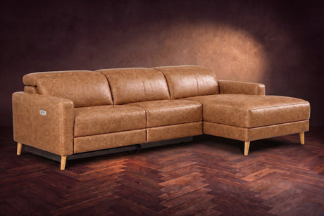 The Elodie sectional on a dark walnut herringbone floor in front of a warm plaster wall, three-quarter view, headrests down and the footrest closed. Shown in Antiqued Caramel, Right chaise.