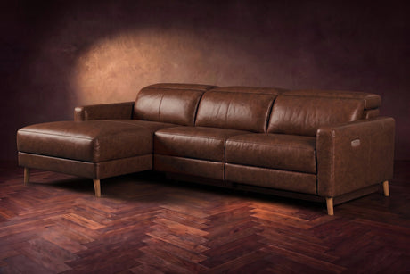 The Elodie sectional on a dark walnut herringbone floor in front of a warm plaster wall, three-quarter view, headrests down and the footrest closed. Shown in Burnished Chestnut, Left chaise.