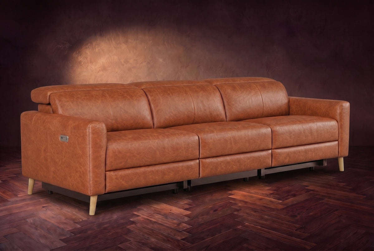 The Elodie sofa on a dark walnut herringbone floor against a plaster wall, three-quarter view, headrests down and footrests closed. Shown in Tuscan Cognac, 3 seat.