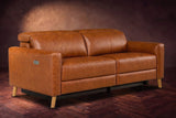 The Elodie loveseat on a dark walnut herringbone floor against a plaster wall, three-quarter view, headrests down and footrests closed. Shown in Tuscan Cognac, Loveseat.