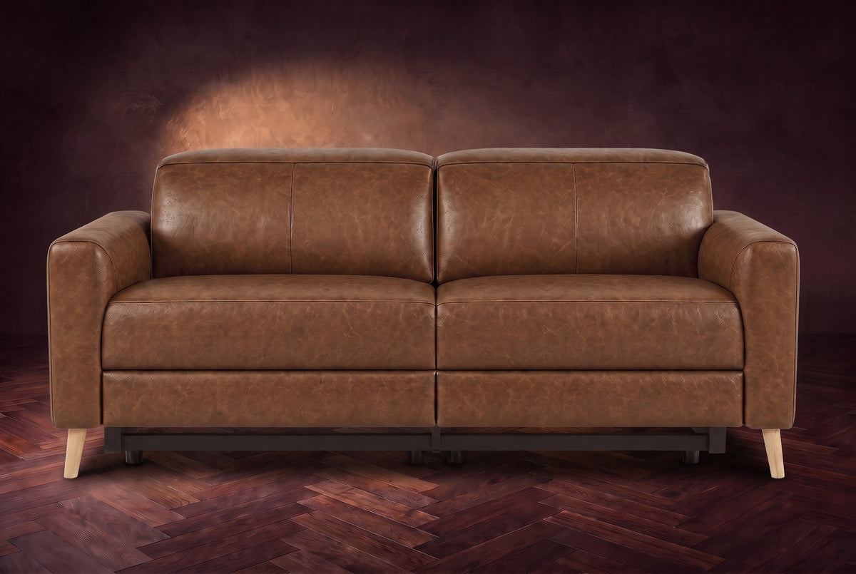 The Elodie loveseat seen from the front on a dark walnut herringbone floor against a plaster wall, headrests down and footrests closed. Shown in Weathered Saddle, Loveseat.