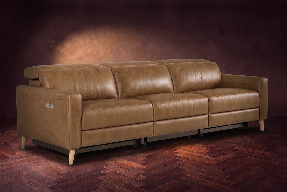 The Elodie sofa on a dark walnut herringbone floor against a plaster wall, three-quarter view, headrests down and footrests closed. Shown in Weathered Saddle, 3 seat.