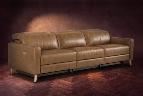 The Elodie sofa on a dark walnut herringbone floor against a plaster wall, three-quarter view, headrests down and footrests closed. Shown in Weathered Saddle, 3 seat.