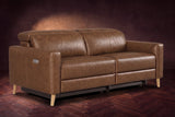 The Elodie loveseat on a dark walnut herringbone floor against a plaster wall, three-quarter view, headrests down and footrests closed. Shown in Weathered Saddle, Loveseat.