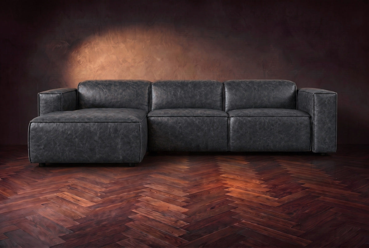 The Emery sectional in Aged Slate distressed leather seen from the front on a dark walnut herringbone floor, headrests down and footrest closed. Shown in Aged Slate, Left chaise.