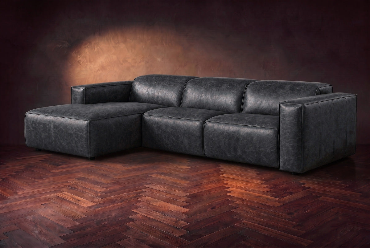 The Emery sectional in Aged Slate distressed leather on a dark walnut herringbone floor against a warm plaster wall, three-quarter view, headrests down and footrest closed. Shown in Aged Slate, Left chaise.