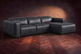 The Emery sectional in Aged Slate distressed leather on a dark walnut herringbone floor against a warm plaster wall, three-quarter view, headrests down and footrest closed. Shown in Aged Slate, Right chaise.