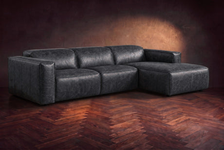 The Emery sectional in Aged Slate distressed leather on a dark walnut herringbone floor against a warm plaster wall, three-quarter view, headrests down and footrest closed. Shown in Aged Slate, Right chaise.