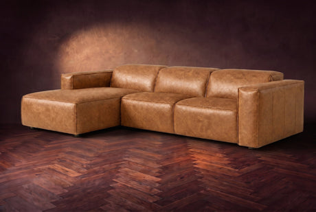 The Emery sectional in Antiqued Caramel distressed leather on a dark walnut herringbone floor against a warm plaster wall, three-quarter view, headrests down and footrest closed. Shown in Antiqued Caramel, Left chaise.