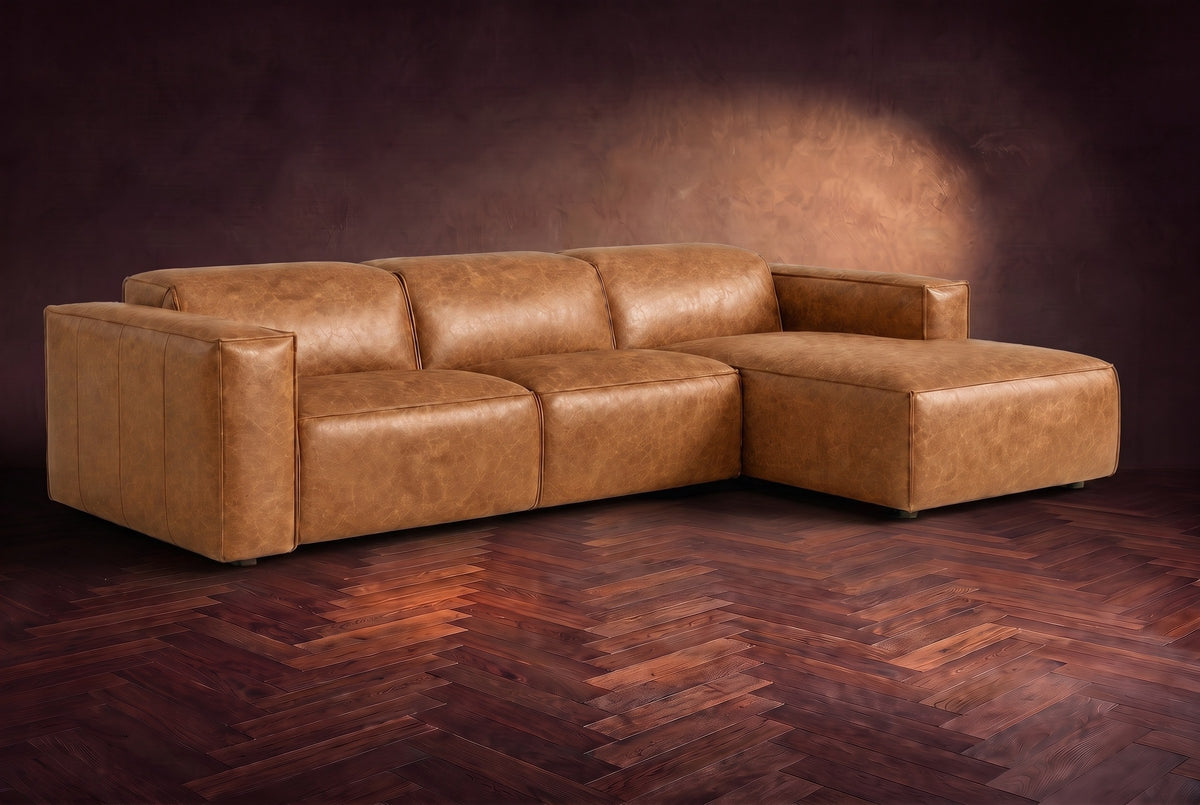 The Emery sectional in Antiqued Caramel distressed leather on a dark walnut herringbone floor against a warm plaster wall, three-quarter view, headrests down and footrest closed. Shown in Antiqued Caramel, Right chaise.