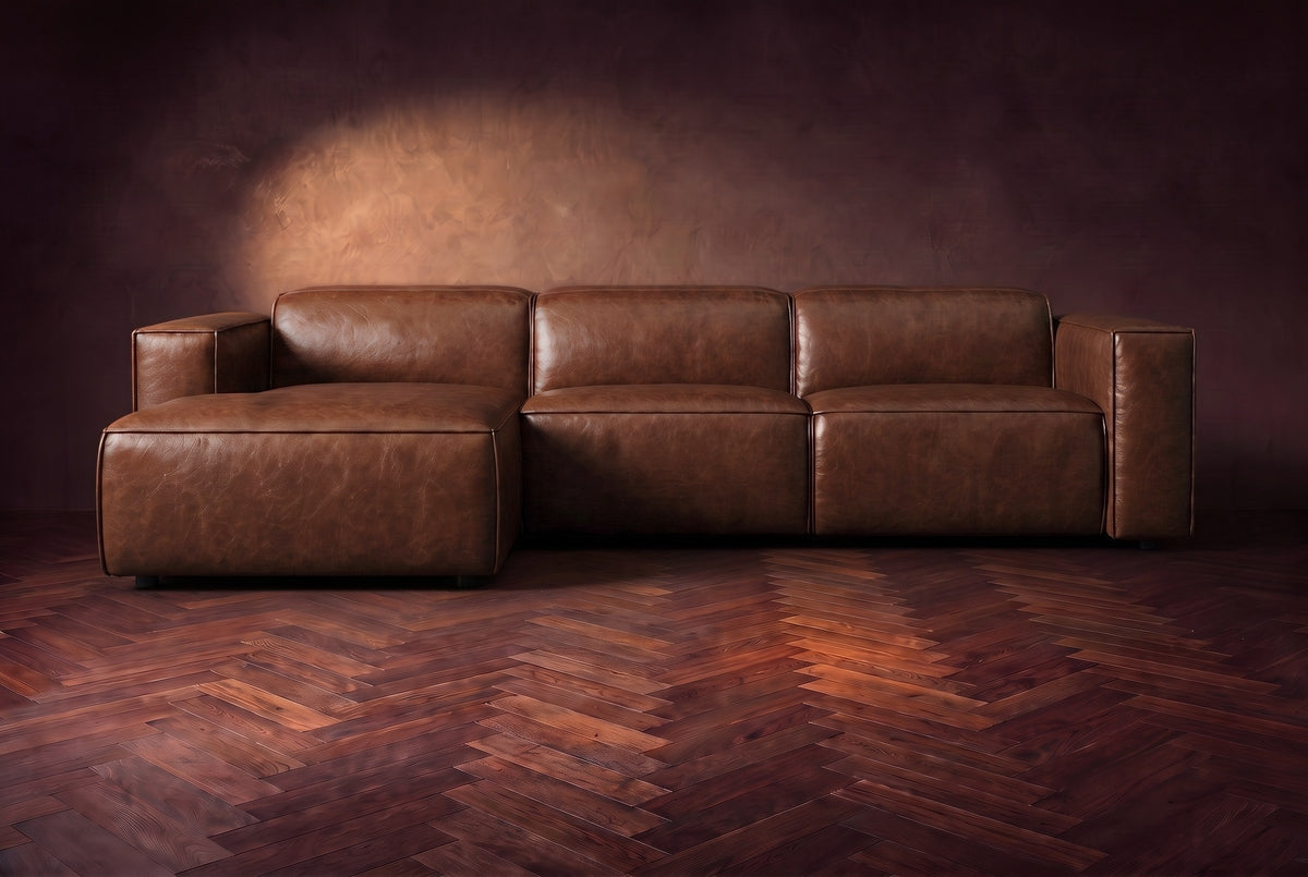 The Emery sectional in Burnished Chestnut distressed leather seen from the front on a dark walnut herringbone floor, headrests down and footrest closed. Shown in Burnished Chestnut, Left chaise.