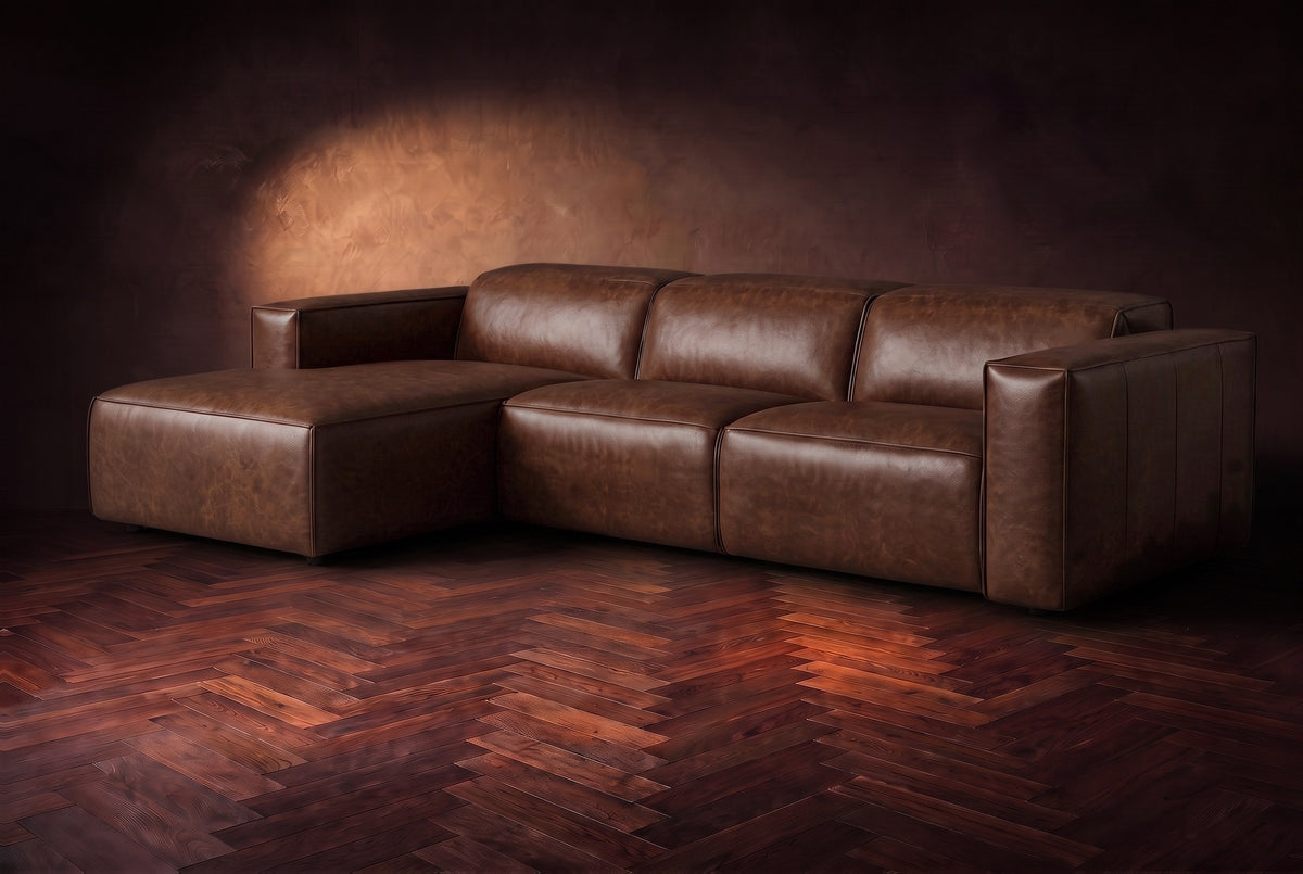 The Emery sectional in Burnished Chestnut distressed leather on a dark walnut herringbone floor against a warm plaster wall, three-quarter view, headrests down and footrest closed. Shown in Burnished Chestnut, Left chaise.