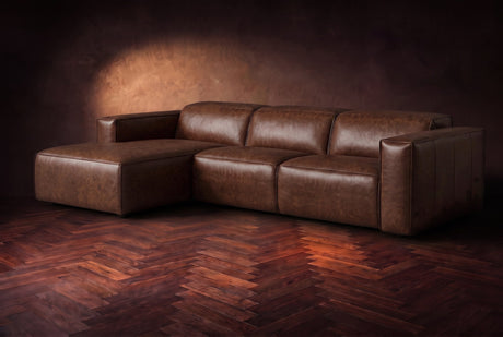 The Emery sectional in Burnished Chestnut distressed leather on a dark walnut herringbone floor against a warm plaster wall, three-quarter view, headrests down and footrest closed. Shown in Burnished Chestnut, Left chaise.