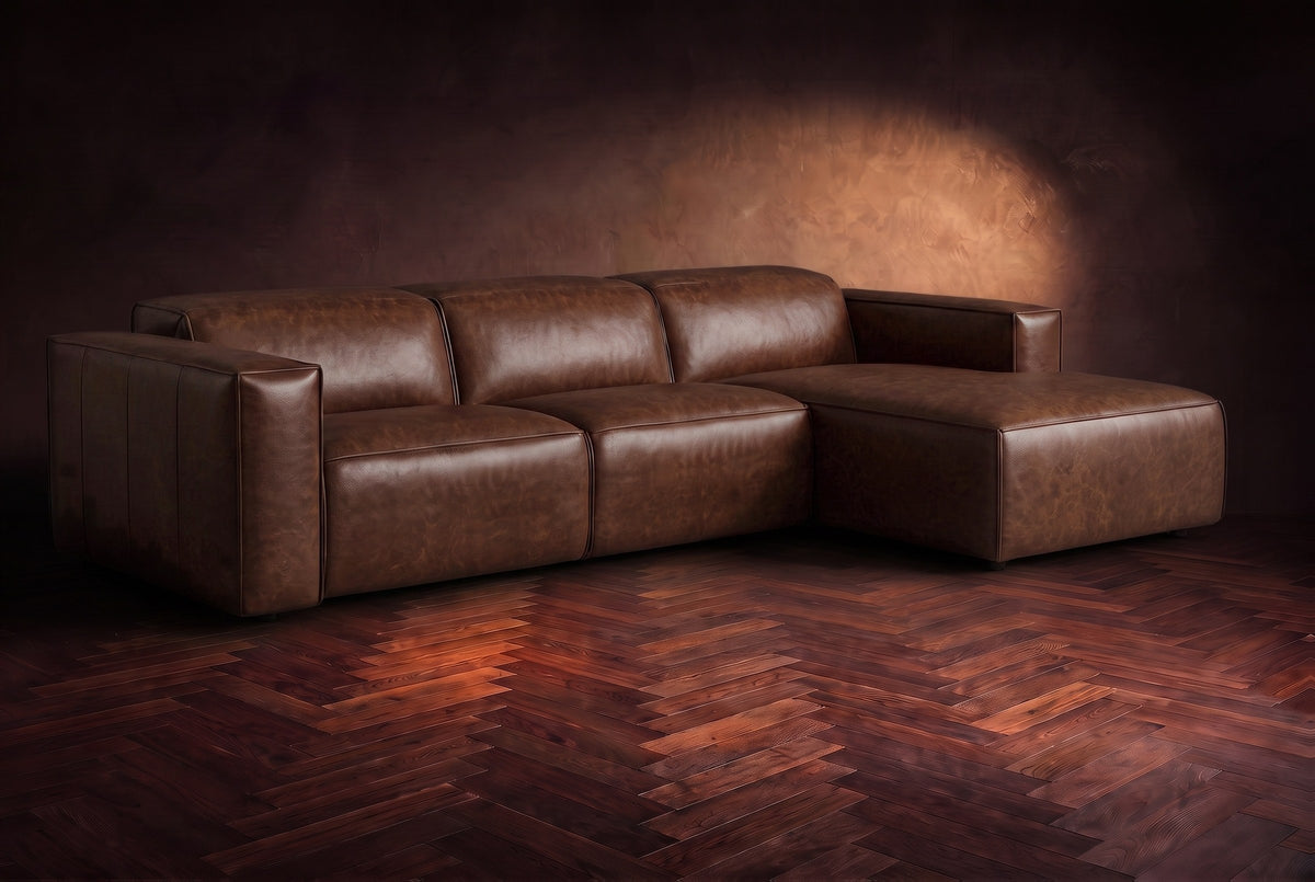 The Emery sectional in Burnished Chestnut distressed leather on a dark walnut herringbone floor against a warm plaster wall, three-quarter view, headrests down and footrest closed. Shown in Burnished Chestnut, Right chaise.