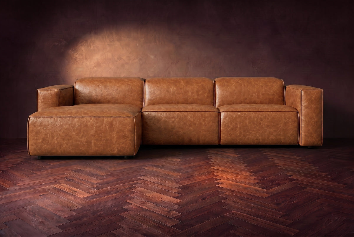 The Emery sectional in Tuscan Cognac distressed leather seen from the front on a dark walnut herringbone floor, headrests down and footrest closed. Shown in Tuscan Cognac, Left chaise.