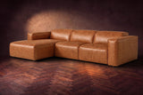 The Emery sectional in Tuscan Cognac distressed leather on a dark walnut herringbone floor against a warm plaster wall, three-quarter view, headrests down and footrest closed. Shown in Tuscan Cognac, Left chaise.