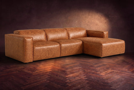 The Emery sectional in Tuscan Cognac distressed leather on a dark walnut herringbone floor against a warm plaster wall, three-quarter view, headrests down and footrest closed. Shown in Tuscan Cognac, Right chaise.