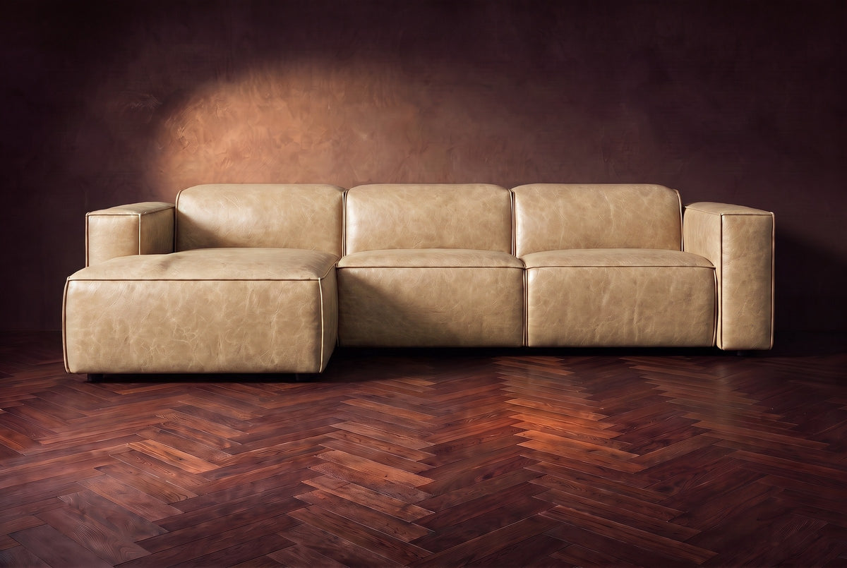 The Emery sectional in Washed Sand distressed leather seen from the front on a dark walnut herringbone floor, headrests down and footrest closed. Shown in Washed Sand, Left chaise.