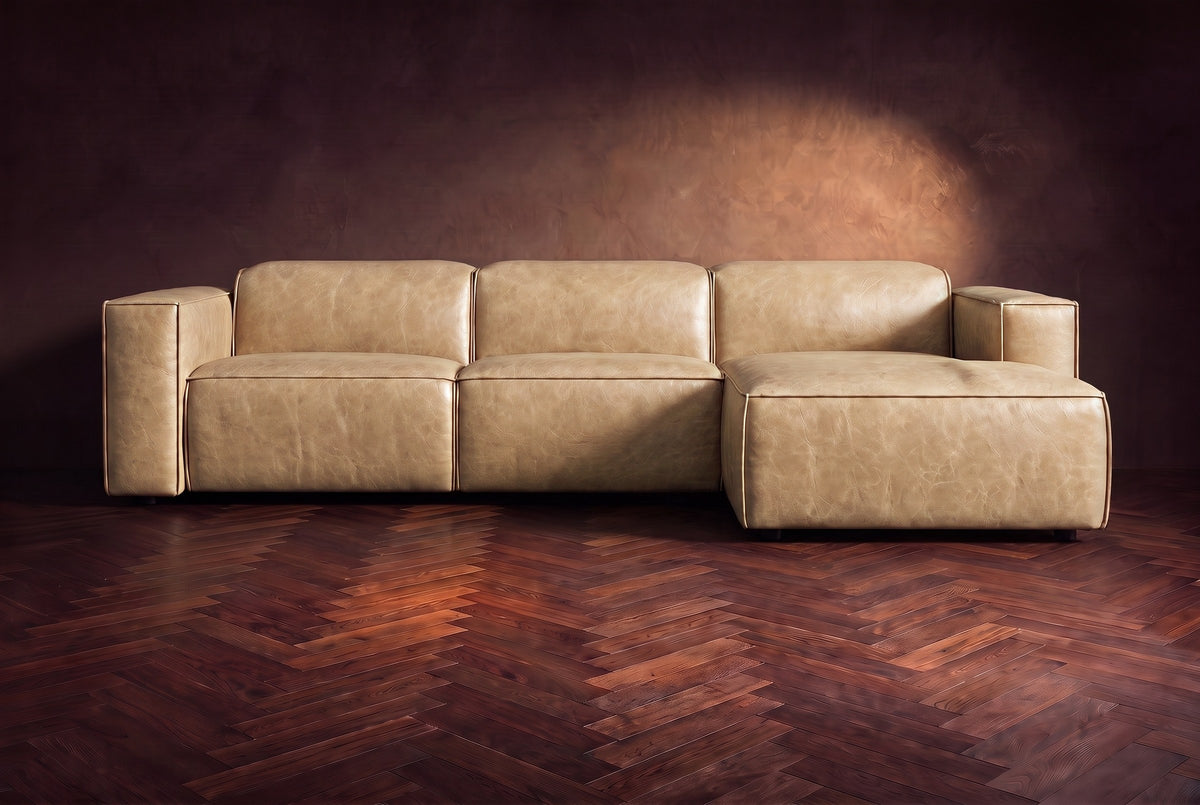 The Emery sectional in Washed Sand distressed leather seen from the front on a dark walnut herringbone floor, headrests down and footrest closed. Shown in Washed Sand, Right chaise.