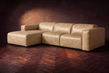 The Emery sectional in Washed Sand distressed leather on a dark walnut herringbone floor against a warm plaster wall, three-quarter view, headrests down and footrest closed. Shown in Washed Sand, Left chaise.