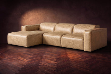 The Emery sectional in Washed Sand distressed leather on a dark walnut herringbone floor against a warm plaster wall, three-quarter view, headrests down and footrest closed. Shown in Washed Sand, Left chaise.