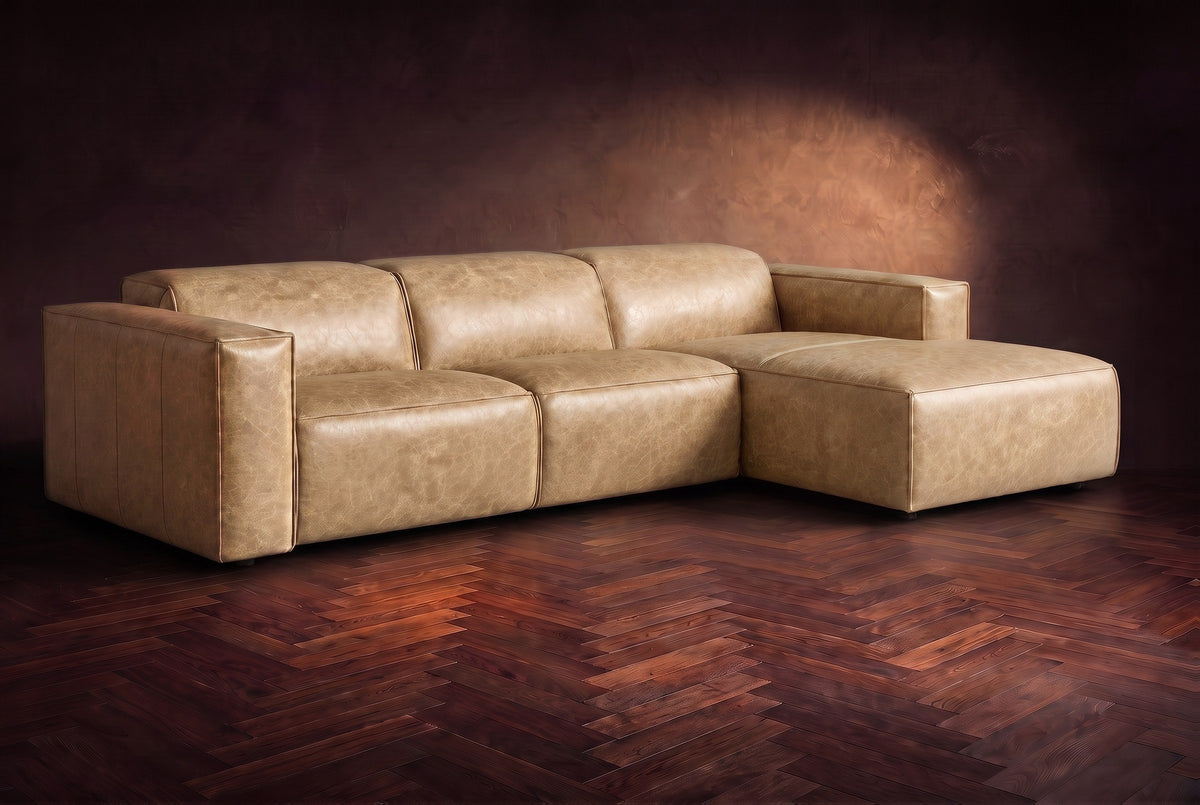 The Emery sectional in Washed Sand distressed leather on a dark walnut herringbone floor against a warm plaster wall, three-quarter view, headrests down and footrest closed. Shown in Washed Sand, Right chaise.