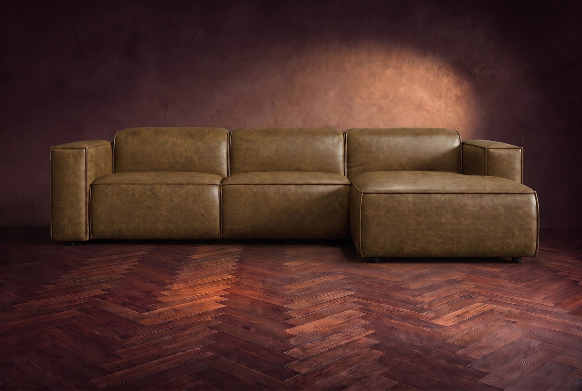 The Emery sectional in Weathered Saddle distressed leather seen from the front on a dark walnut herringbone floor, headrests down and footrest closed. Shown in Weathered Saddle, Right chaise.