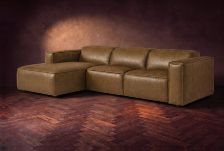 The Emery sectional in Weathered Saddle distressed leather on a dark walnut herringbone floor against a warm plaster wall, three-quarter view, headrests down and footrest closed. Shown in Weathered Saddle, Left chaise.