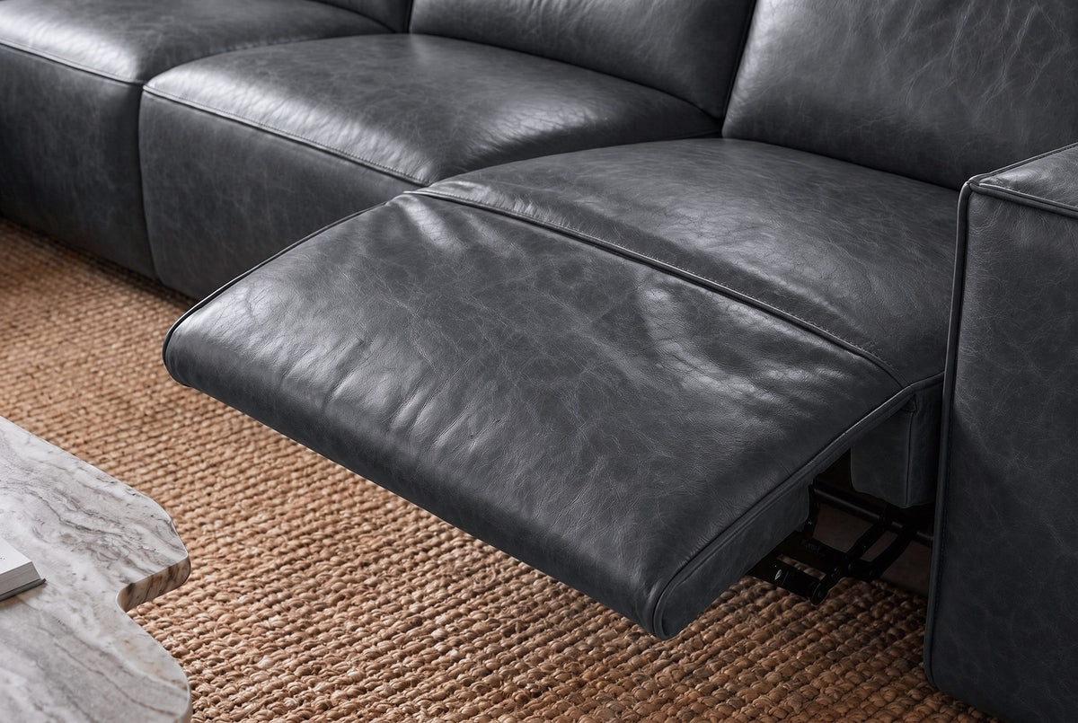 Close-up of an Emery recliner beside its box arm, the one-piece footrest out over its black steel mechanism and the closed seat beside it in line, on a woven jute rug. Shown in Aged Slate.