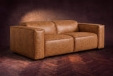 The Emery loveseat in Antiqued Caramel distressed full leather on a dark herringbone floor, headrests down and footrests closed. Shown in Antiqued Caramel, Loveseat.