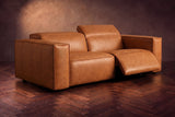 The Emery loveseat in Tuscan Cognac on a dark herringbone floor, the right-hand recliner open with its one-piece footrest out and its headrest raised. Shown in Tuscan Cognac, Loveseat.