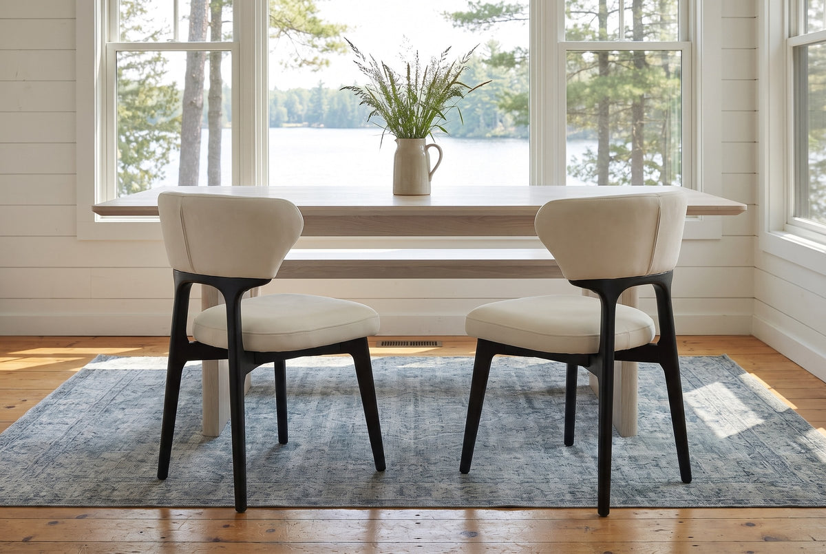 Two Fosdinovo dining chairs on a blue-grey rug in front of a pale oak table in a Muskoka cottage with a window onto the lake