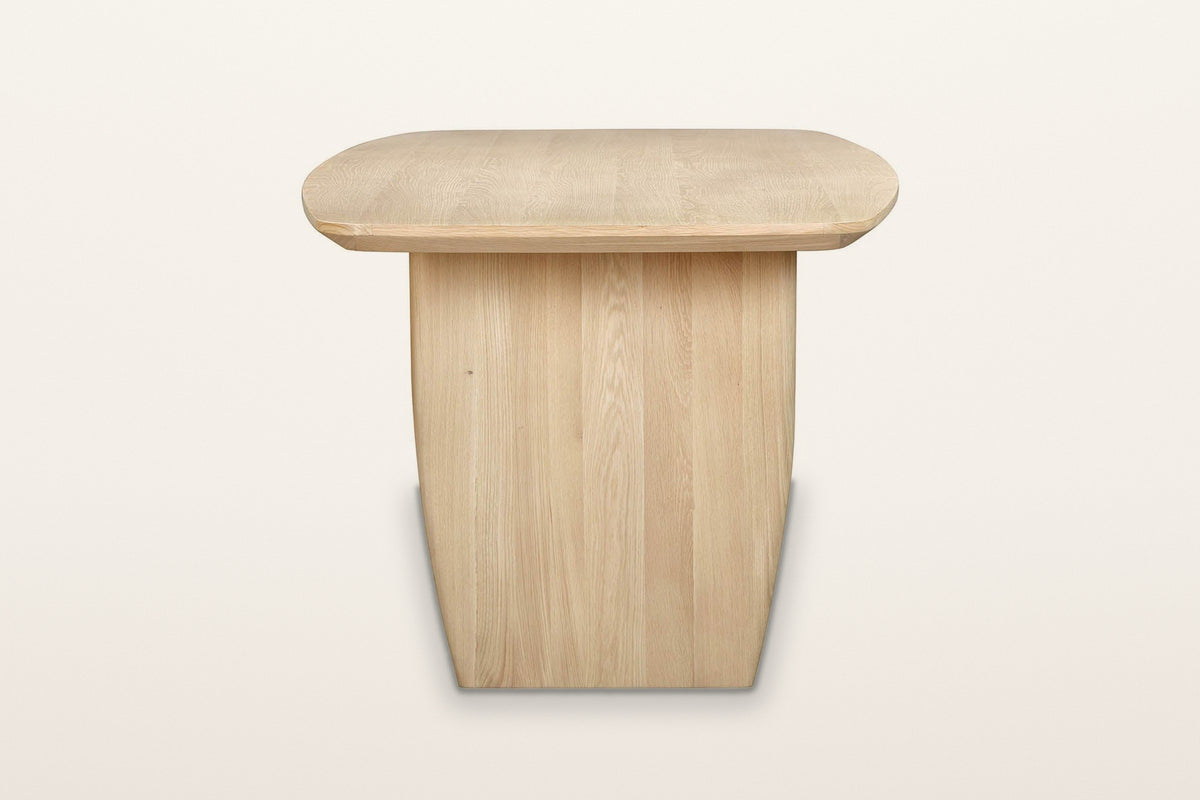 Frontino dining table in Biscotti Oak, end view on a warm white studio backdrop: one wide slab leg under the rounded end of t