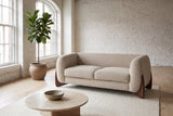 Greccio boucle loveseat in almond in a white-brick loft with arched windows, a fig tree and a round travertine table